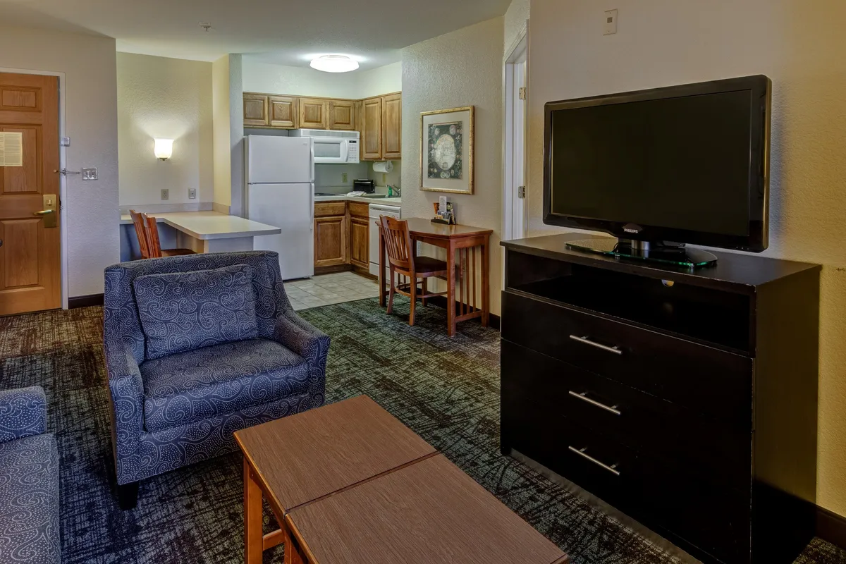 The image showcases a bright and inviting open-concept living space in the condo, featuring a comfortable sofa, a stylish dining table, a modern kitchen area, a flat-screen television, and a chest of drawers. The area is well-lit and thoughtfully arranged for both relaxation and socializing, capturing a cozy, homey mood.