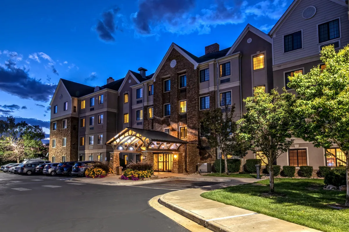 The exterior shot highlights the property's well-lit entrance awning, landscaped gardens, and parked cars. The warm glow from the windows and welcoming atmosphere set the scene for a family's return after a day of exploring, emphasizing comfort and convenience.