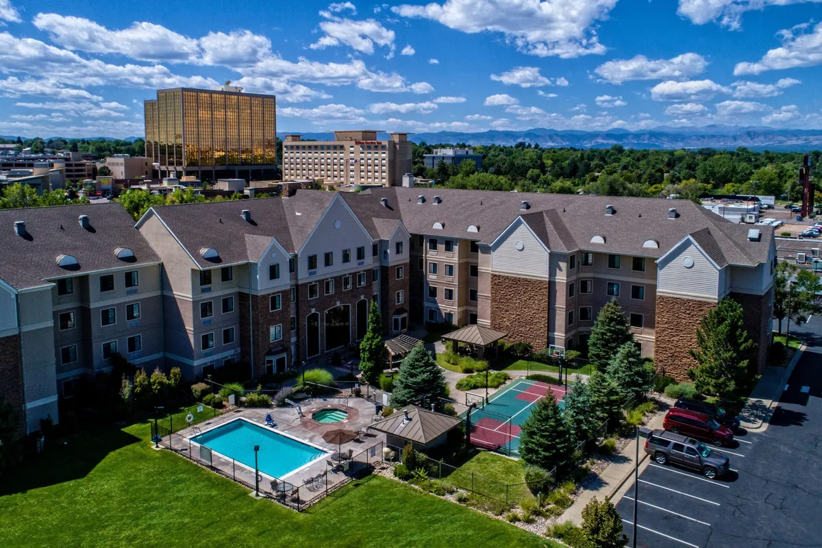 A vibrant exterior view featuring the outdoor swimming pool, adjacent hot tub, tennis courts, and lush landscaped gardens. Guests can be seen enjoying the pool, sunbathing, and relaxing in the hot tub, with the building facade in the background suggesting a communal, resort-like environment.