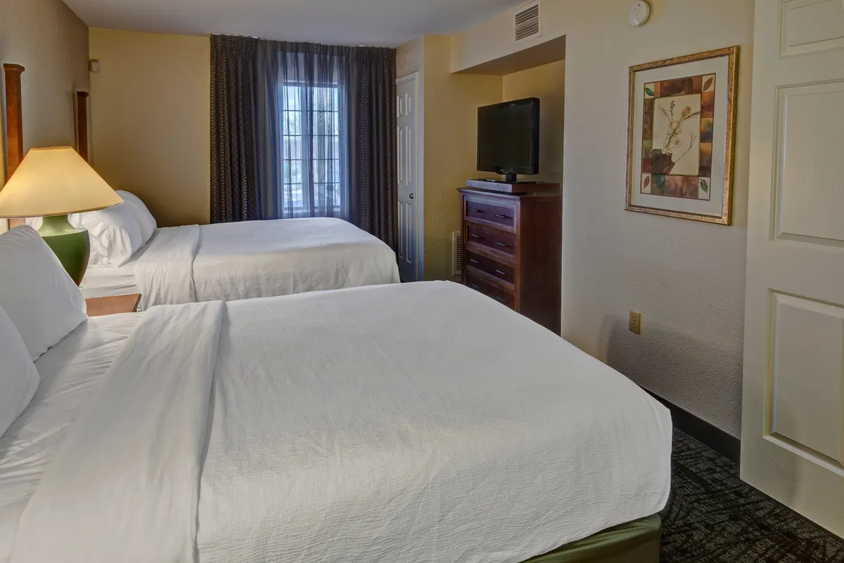 A well-lit bedroom featuring two queen beds dressed in crisp linens, a window with soft curtains allowing natural light, a TV atop a dresser for in-room entertainment, and tasteful framed artwork that adds warmth to the ambiance.