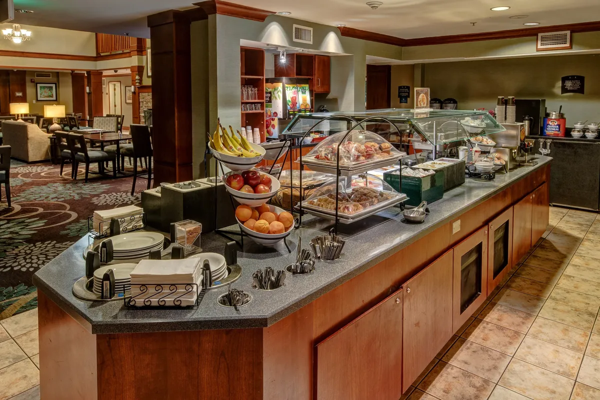 The image features a welcoming breakfast buffet with a diverse selection of fresh fruits and elegantly arranged pastries. Comfortable seating is visible in the adjacent dining area, inviting guests to enjoy their meals or coffee in a communal setting.