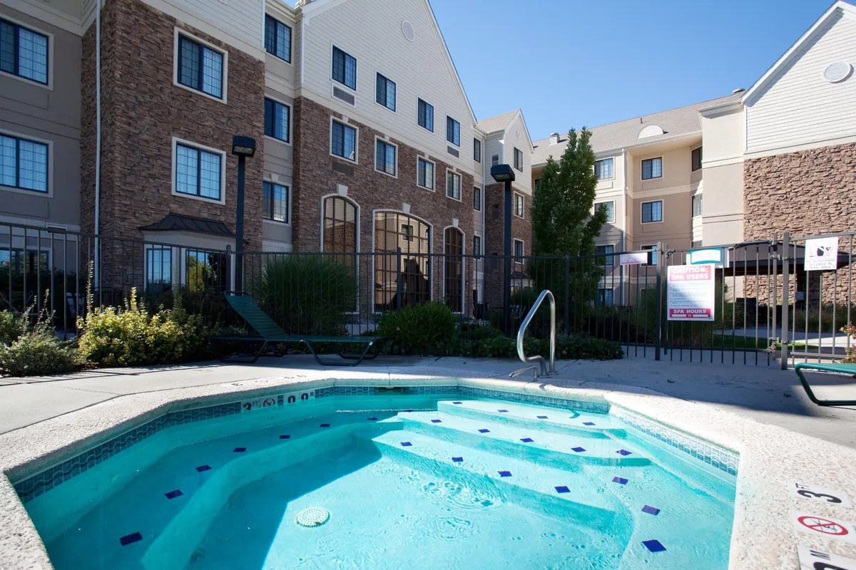 A close-up exterior shot of the hot tub area, surrounded by well-maintained landscaping and the building's inviting facade. The scene is tranquil yet communal, with guests enjoying a soak at sunset and others socializing nearby.