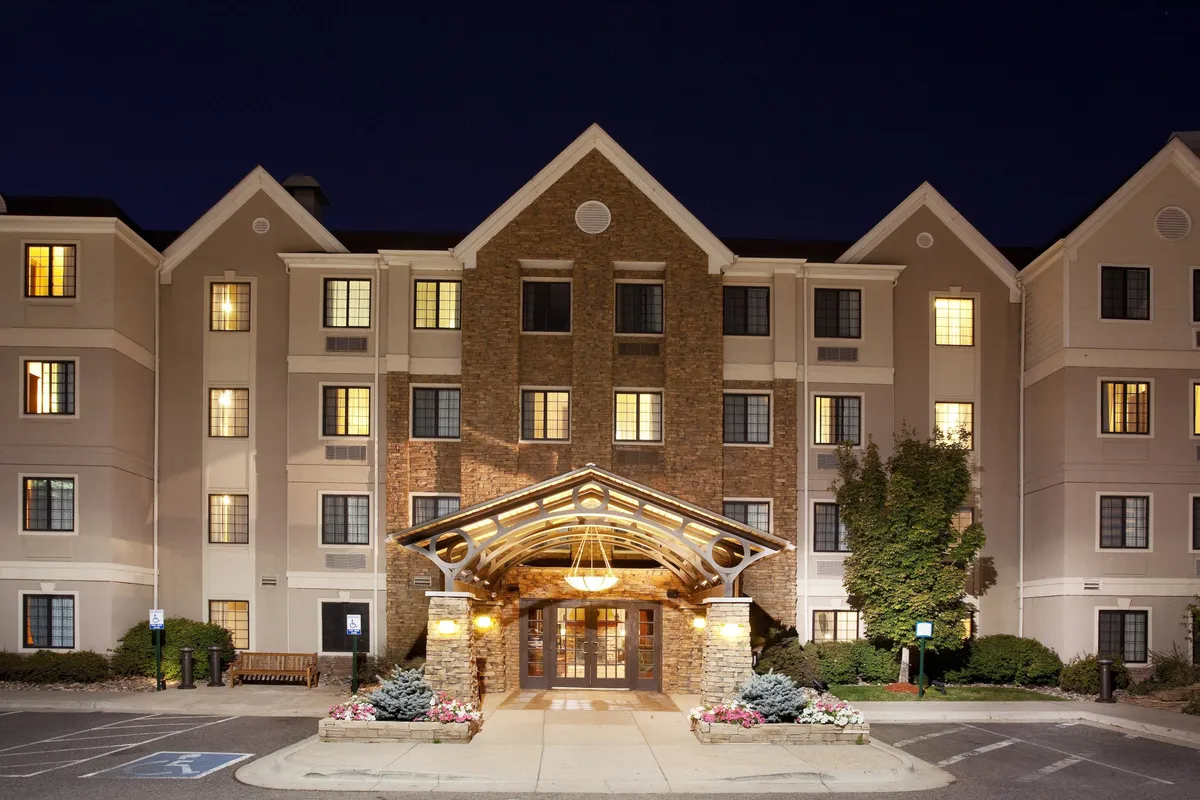 The image captures the exterior of the property, showcasing the welcoming entrance canopy, attractive stone facade, and warmly illuminated windows in the evening. Guests are seen checking in, relaxing in the outdoor seating area, and casually exploring the area around the entrance, emphasizing the property's inviting atmosphere and easy accessibility.